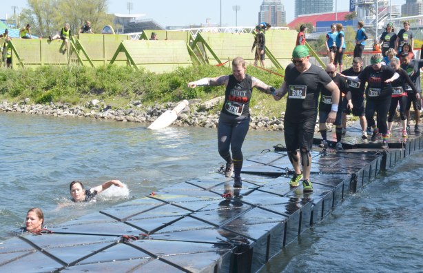 Two runners are holding hands as they cross a very tippy and wobbly floating bridge, many runners are behind them, waiting their turn to cross 