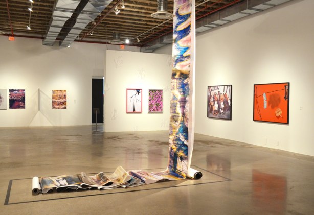 pictures on an art gallery wall.  In the middle of the room is a large roll of photographic paper that has been developed with streaks of colour.  It hangs from the ceiling and lays on the floor. 