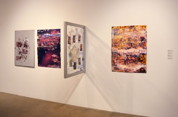 Four pictures on an art gallery wall, all abstract.  One of them protrudes from the wall at a 90 degree angle. 