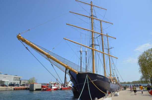 The tall shil 'Caledonia' is moored in the foreground.  Behind and to the left are red fire boats, to the right is a walkway beside the lake on which people are walking and cycling. 