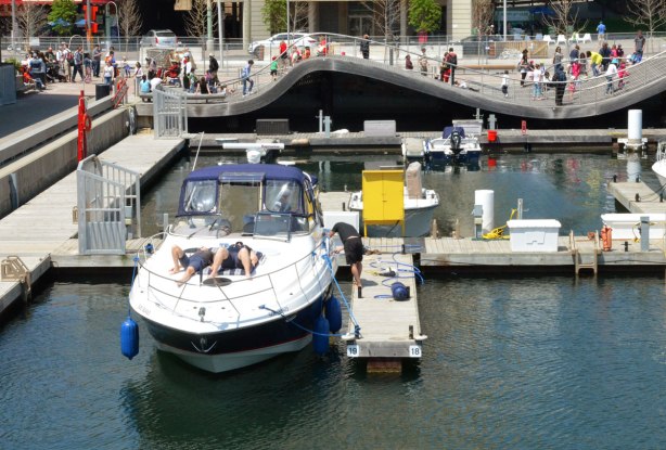 Two people are lying on the deck of a large boat that is moored at the waterfront, in the background is the Rees wave deck, a wavy sidewalk, on which a lot of people are walking 