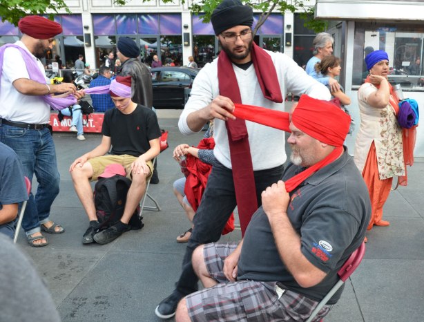 Two men sitting in chairs while having their heads wrapped in a turban, one purple and one orange. 