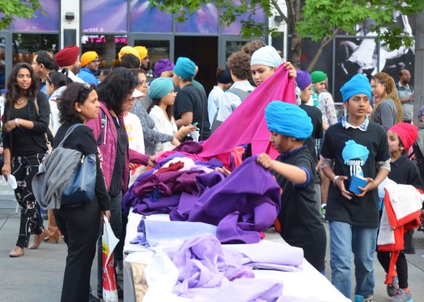 People in turbans are at a table sorting and folding purple and pink and blue fabric that is available for making turbans with. 