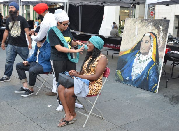 A black woman with long hair is having her head wrapped in a turquoise turban