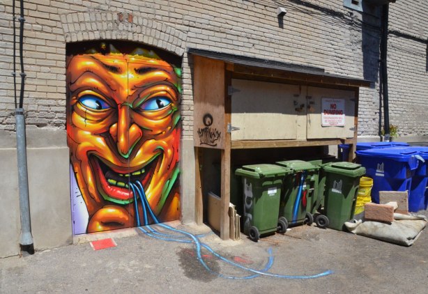 graffiti on a door of a large grotesque man's face with open mouth and what looks like blue ribbons coming out of his mouth, the blue ribbons continue on the concrete of the lane behind the door, and go towards the garbage bins beside the door.