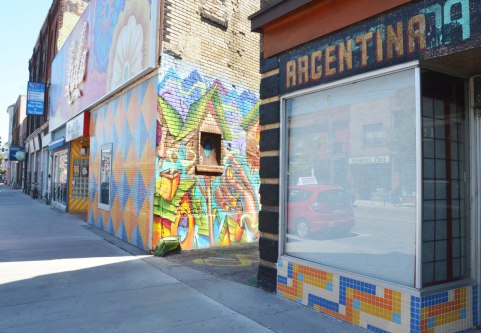 street scene on Dundas West, sidewalk and store fronts, with mural in the alley just showing betweenthe Lula lounge with its orange, blue and white tiled front and a store with the sign Argentina on it.