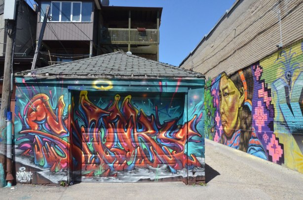 a garage door covered with a street art piece signed by smoky, a mural in the lane beside the garage is also visible