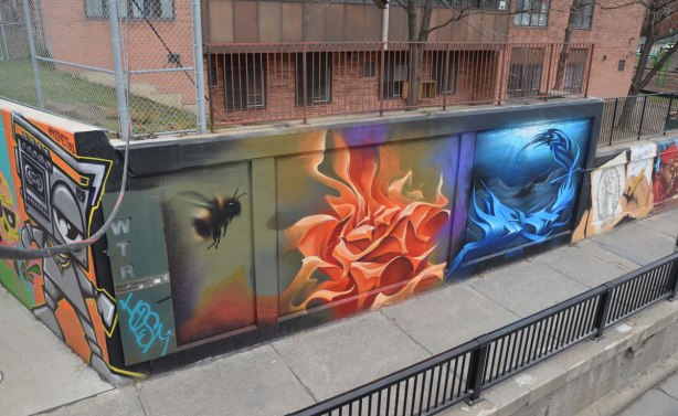 Street art under a railway overpass as seen from on top of the bridge.  A painting of a bumblebee flying towards a bright yellow and orange flower as well as a painting that is done is blues can be seen. 