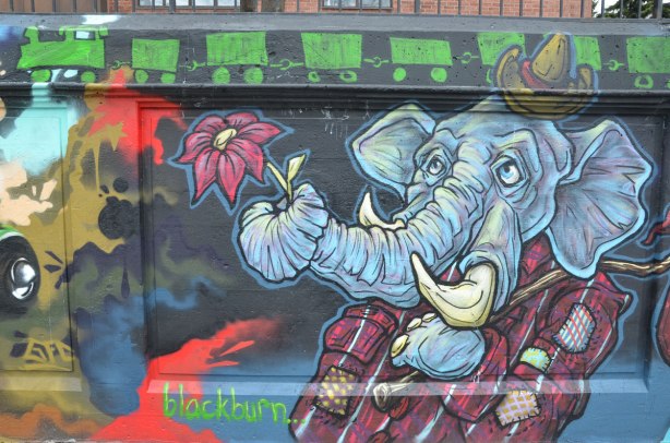 street art painting of an elephant standing upright, only upper part shown, wearing a plaid jacket, carrying a cane over his shoulder, a small hat on his head, and holding a flower in his trunk.   A green train runs across the top of the painting.   signed by blackburn