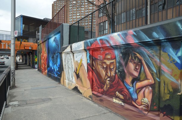 looking along a sidewalk that runs under a railway bridge.  Along the right hand side are a series of graffiti paintings. 