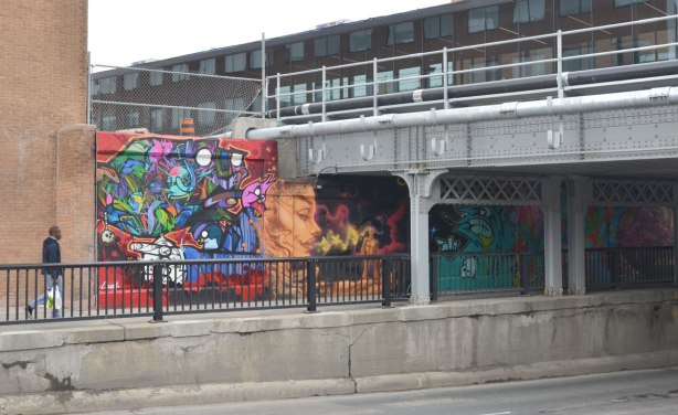 Man walking along a sidewalk towards a railway bridge.  One side of the underpass has been painted with street art pictures. 
