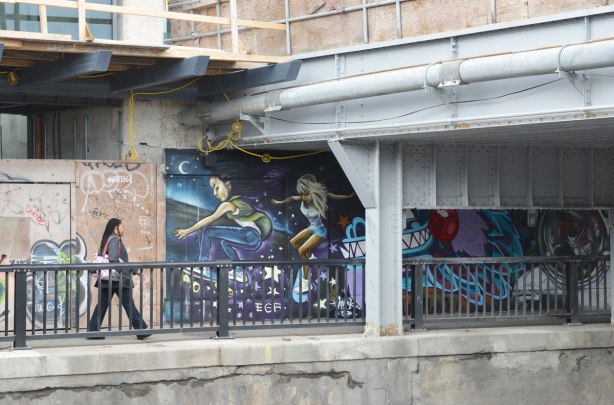 woman walking along a sidewalk under a railway bridge.  There is a graffiti picture of two people skateboarding beside her. 