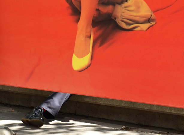 a man's leg and black boot stick out from under a picture of a woman's leg with a yellow shoe, on a bright orange background. A man is sitting on a ledge behind the photo.