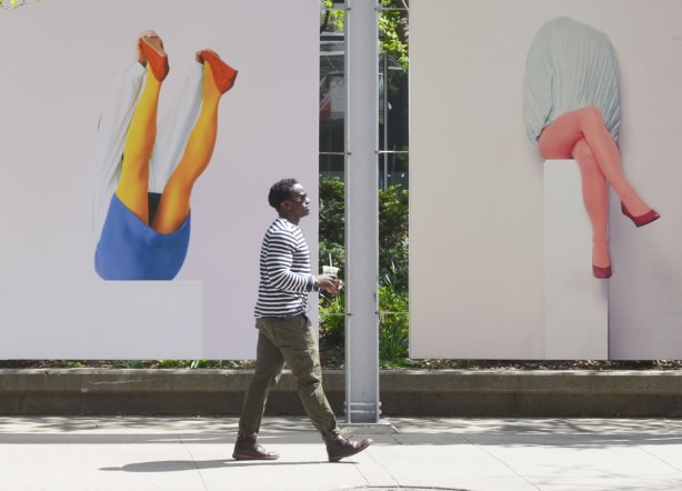 A man in brown pants and a brown and white striped shirt is holding a drink in his hand as he walks past two large pictures of legs.