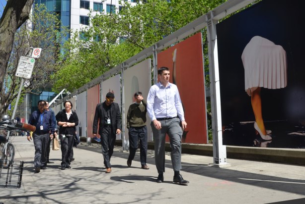 Six people walking on a sidewalk. They are walking past a row of large pictures of legs in various strange poses on bodies with no upper parts.