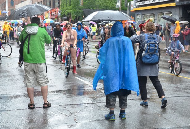 A group of women on bikes are riding in a dyke march, pride week. The march has temporarilty stopped and three photographers are standing in front of the riders taking their picture. The woman in front is topless and she has drawn a picture of a bike on her breasts.