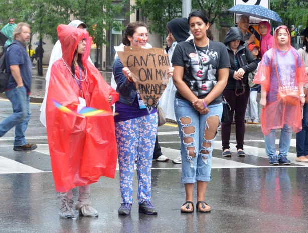 Three young women standing in the rain as the watch a Dyke march parade. One is wearing a red poncho and one is holding a sign that says " Can't rain on my parade"