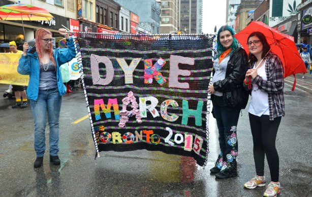 Three women are holding a large banner that says 'Dyke March Toronto 2015' that has been crocheted and sewn together.
