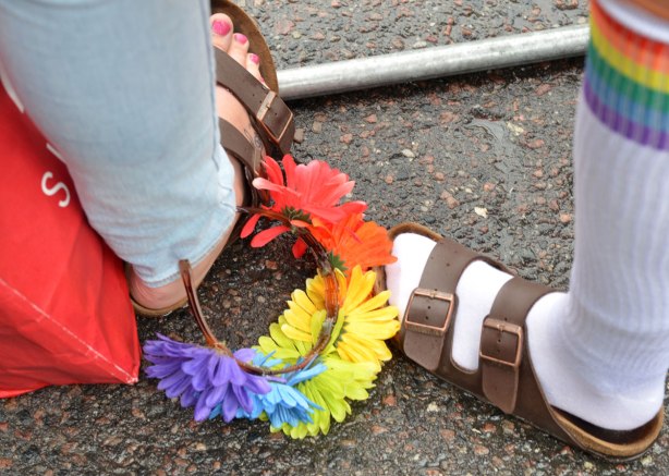 A flower head band has fallen to the ground and is beside two people's feet, both of whom are wearing sandals. 