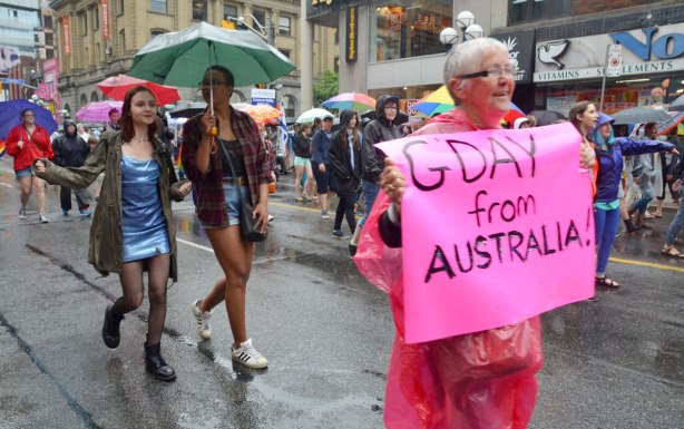 pride week, dyke march, a grey haired woman is carrying a pink sign that says 'G'day from Australia' and behind her is a couple of young women sharing a gren umbrella