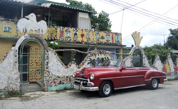 a Cuban house behind a wall that is covered with tile mosaic.  An old red car is parked in front of the building. 
