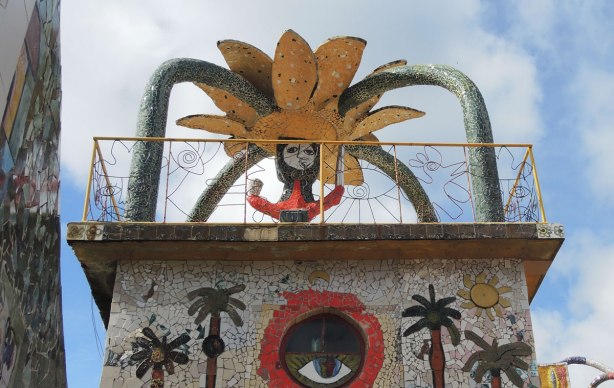 The words 'sea feliz y ria' painted on tiles around the bottom of a balcony. A tile covered statue of a red person with a large flower on their head is also in the picture. The balcony is surround by a wire fence, with faces made of metal open work in the railing too.
