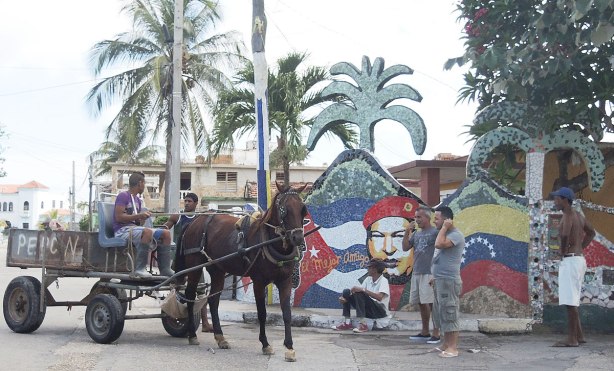 A horse drawn wagon and some men standing around on a corner.  A mural made of ceramic tiles is behind them including a life size cut out of a palm tree 