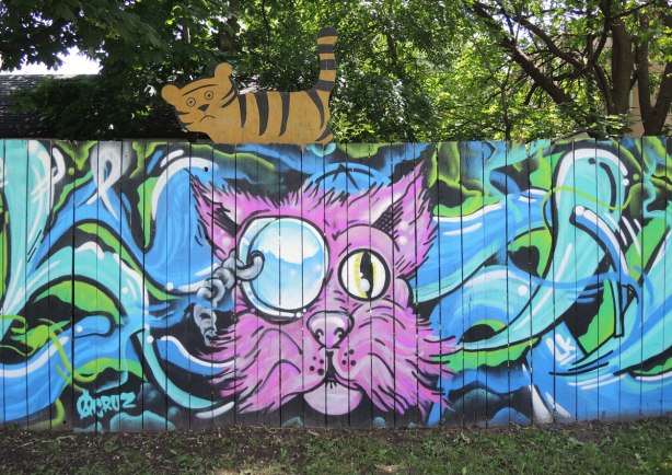 Street art painting of a pink cat head on a fence with a cutout brown and black cat above it. 