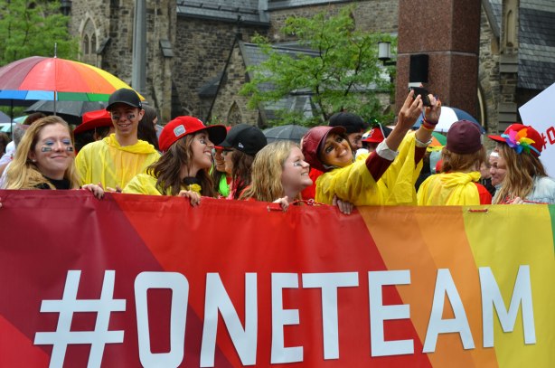 A group of people holding a large rainbow coloured banner that says #oneteam.  Some of them are taking selfies 