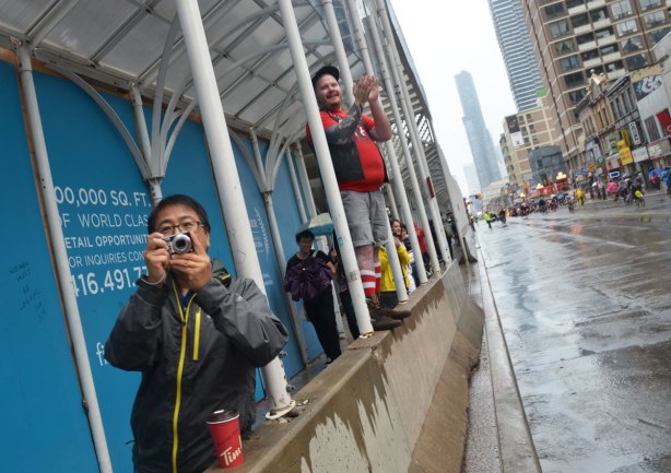Two men watching a parade on a rainy afternoon. One is holding a camera and is standing behind a concrete barricade. The other man is standing on the barricade and cheering and clapping.