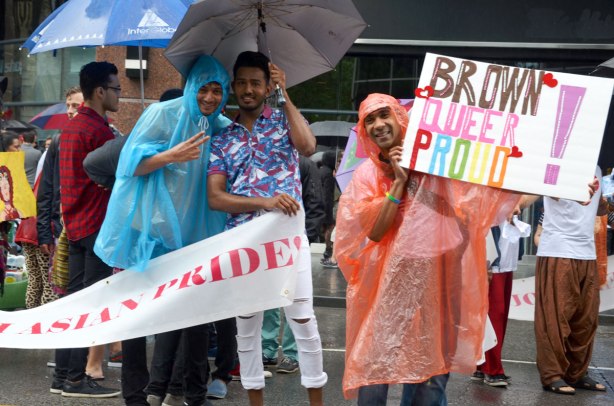 people at a pride parade in the rain - three Asain guys, two under an umbrella, and a third wearing a salmon coloured rain poncho and holding up a sign that says "brown queer proud'