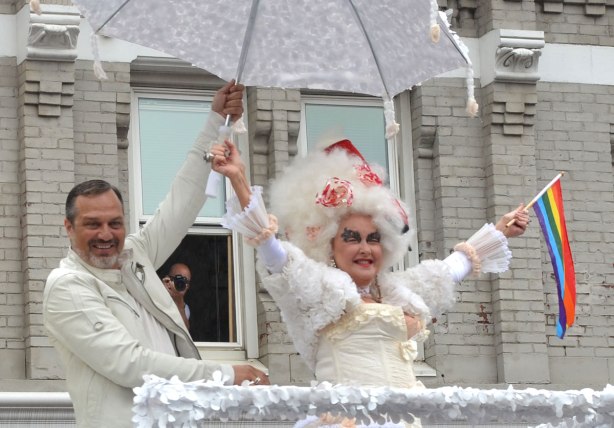 people at a pride parade on a rainy day - Cyndi Lauper is wearing a white frilly dress and hat as she rides on top of a float for the musical KInky Boots.  In the background a photographer leans out the window of a building to get a better view of the parade. 