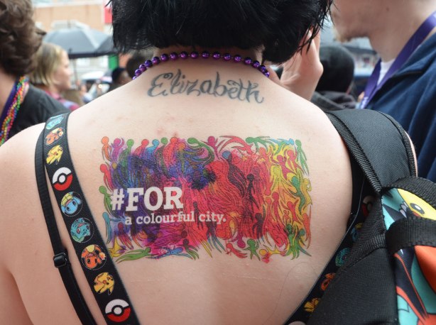 people at a pride parade on a rainy day - the back of a woman with short black hair.  She has a tattoo across the back of her neck that says Elizabth as well as a temporary colourful tattoo that includes the words #for a colourful city