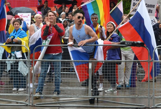 people at a pride parade on a rainy day - one of the parade marchers, while holding a French flag, stops to lift his leg over the barricades
