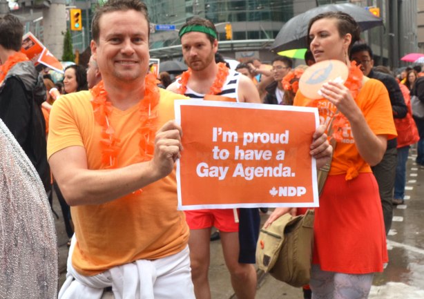 people at a pride parade in the rain - a man in an orange Tshirt holding a sign that says "I'm proud to have a gay agenda, NDP"