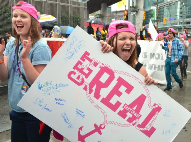 A young woman making a face at the camera as she holds a sign that says "get real" - people at a pride parade in the rain