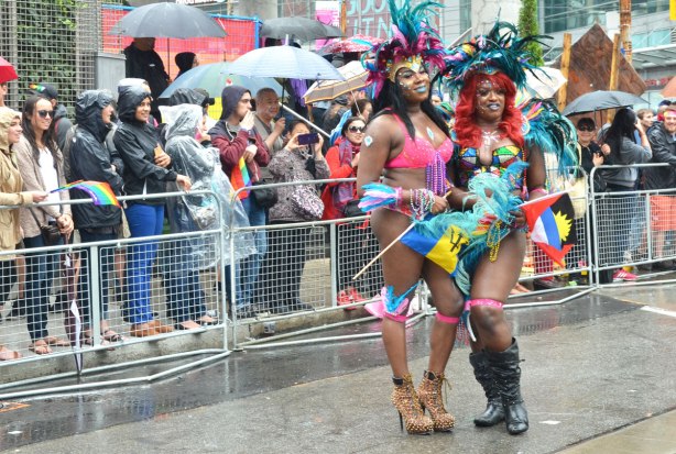 people at a pride parade in the rain - two guys in drag with high heeled boots and wigs, with lots of feathers on their heads, are posing in front of a crowd who are behind metal barricades. 