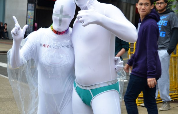 people at a pride parade on a rainy day - the #HIVanonymous characters in complete white costume stop to pose for a picture in front of group that is preaching anti-gay messages