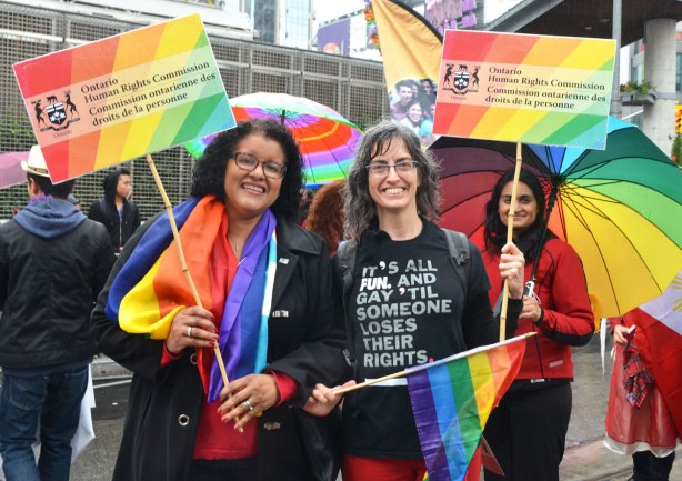people at a pride parade in the rain - some women holding signs promoting the Human RIghts Commission.  they are holding rainbow flags as well 