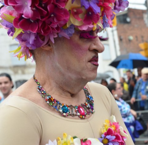 people at a pride parade on a rainy day - an older man in a flowery wig and flowery top along with a bulky necklace stands in front of a crowd, close up shot of head and shoulders