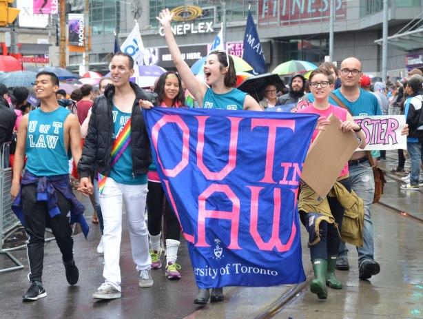 A group from the University of Toronto marches in a pride parade.  They have a large blue banner with pink lettering that says Out in Law. 