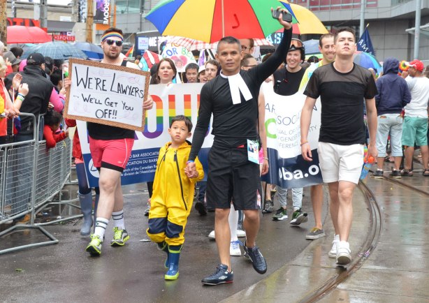 A group representing lawyers walks in the pride parade including a boy in a yellow rain coat.  One of the men is carrying a sign that says We're lawyers, we'll get you off. 