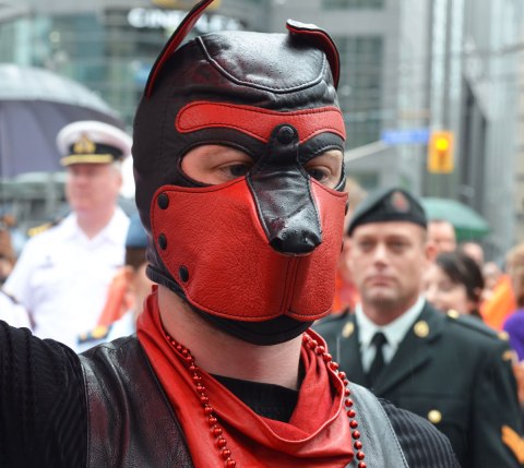people at a pride parade in the rain - close up of a man in a red and black leather mask that covers his whole head, behind him and slightly blurry are two soldiers, navy guy in white on one side and army guy in greenish uniform on the other side 