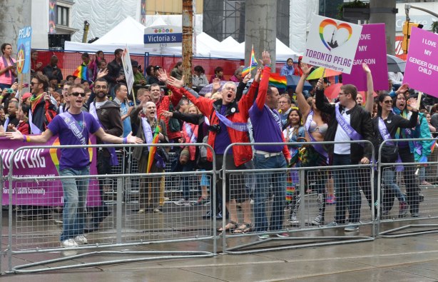 A crowd of marchers cheer -  people at a pride parade on a rainy day -