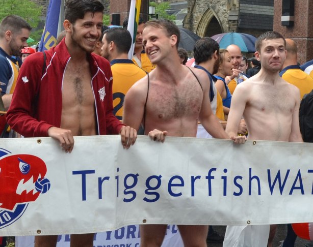 people at a pride parade on a rainy day - a couple of members of the triggerfish waterpolo team stand behind a banner with their team name on it.  They are wearing bathing suits only