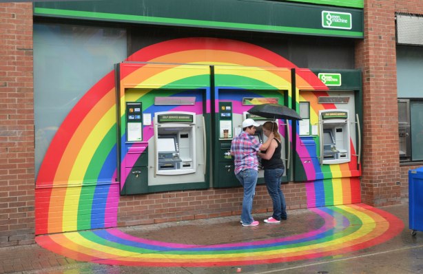 A couple is at an ATM on the side of the TD Canada Trust bank. A large rainbow has been painted around it for pride week.