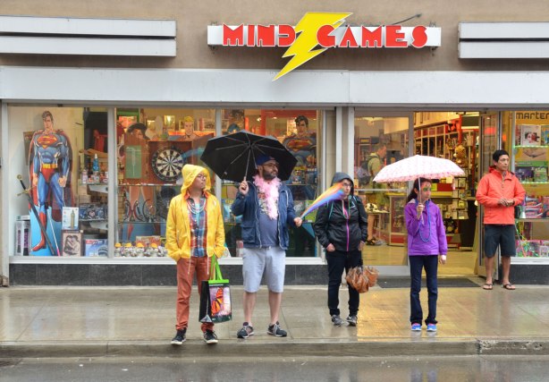 A group of people standing on a sidewalk in the rain as they watch a parade. They are standing in front of a Mind Games store .