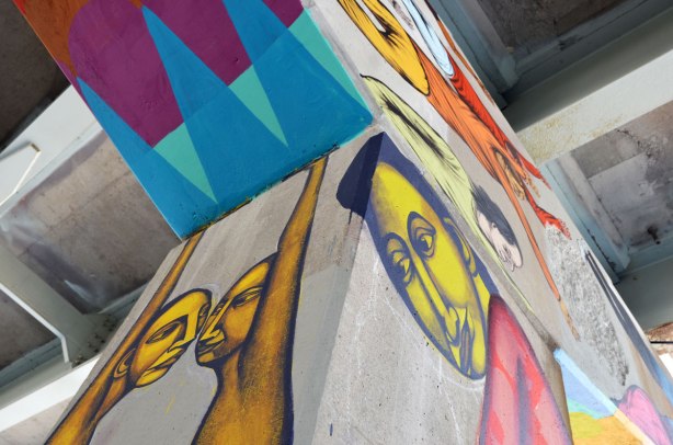 on two sides of a concrete pillar there are paintings of people. One has two nose to nose with both arms raised to the top of the pillar such that it looks like they are holding up the upper horizontal part of the concrete support. On the other side of the pillar is a man with bent head who looks like he is carrying the weight of the road above on his shoulders. There are also some figures painted on the horizontal part of the support.