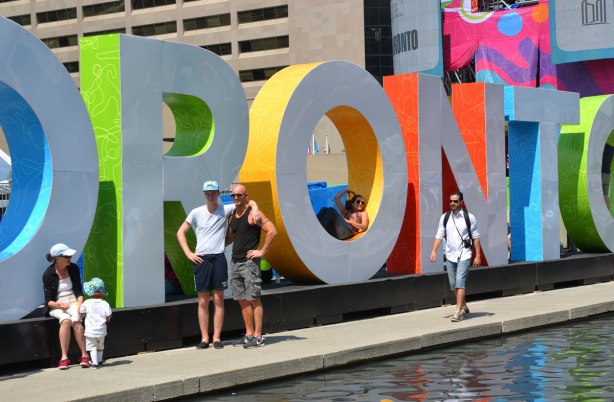 large three dimensional block capital letters that spell Toronto installed alongside the pool fountain in Nathan Phillips Square -  a couple stands in front of it, a man with a camera walks past it 