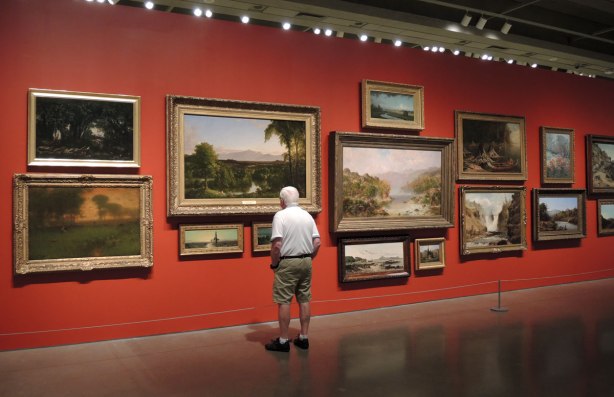 A man is looking at a wall of paintings. The wall is painted red. It is in an art gallery
